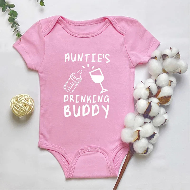 Pink baby onesie with 'Auntie's Drinking Buddy' text and graphics on a light background with cotton and yarn.