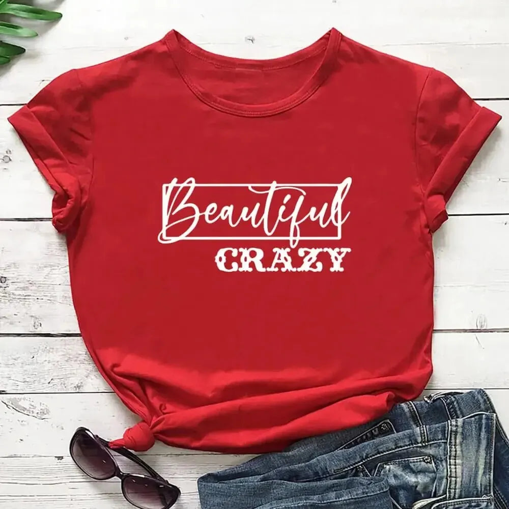 Red t-shirt with 'Beautiful Crazy' text on a wooden surface with sunglasses and jeans.