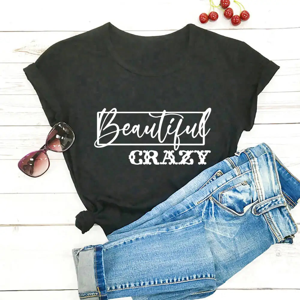Black t-shirt with 'Beautiful Crazy' text, sunglasses, and denim shorts on a wooden surface.