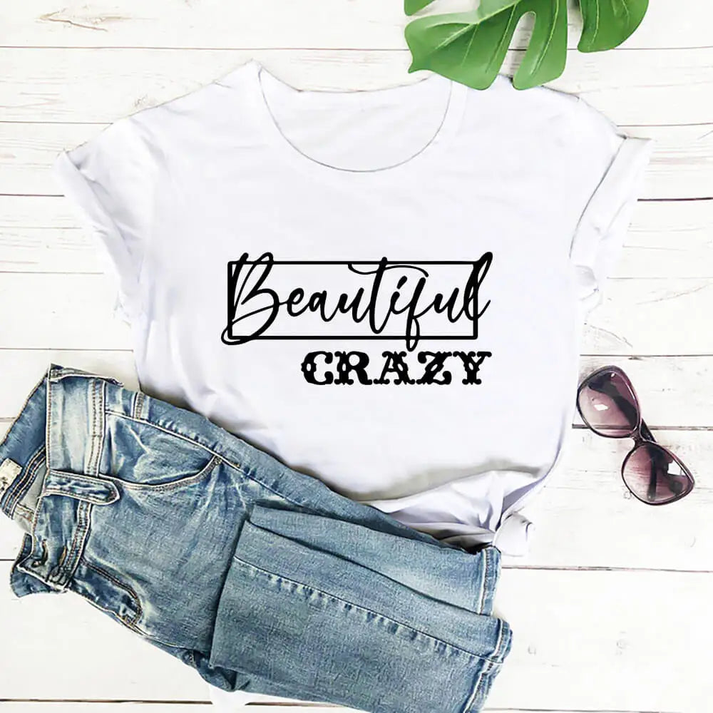 White t-shirt with 'Beautiful Crazy' text, paired with blue jeans and sunglasses on a light wooden surface.