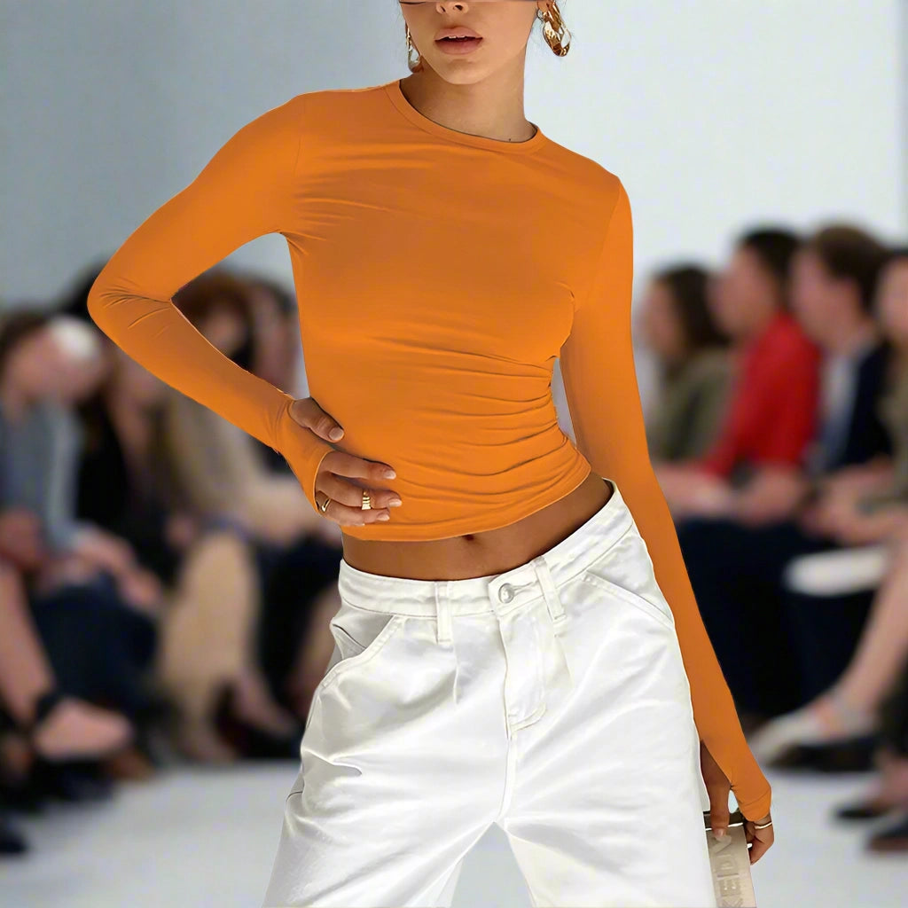Model wearing an orange long-sleeve top and white pants on a runway with an audience.